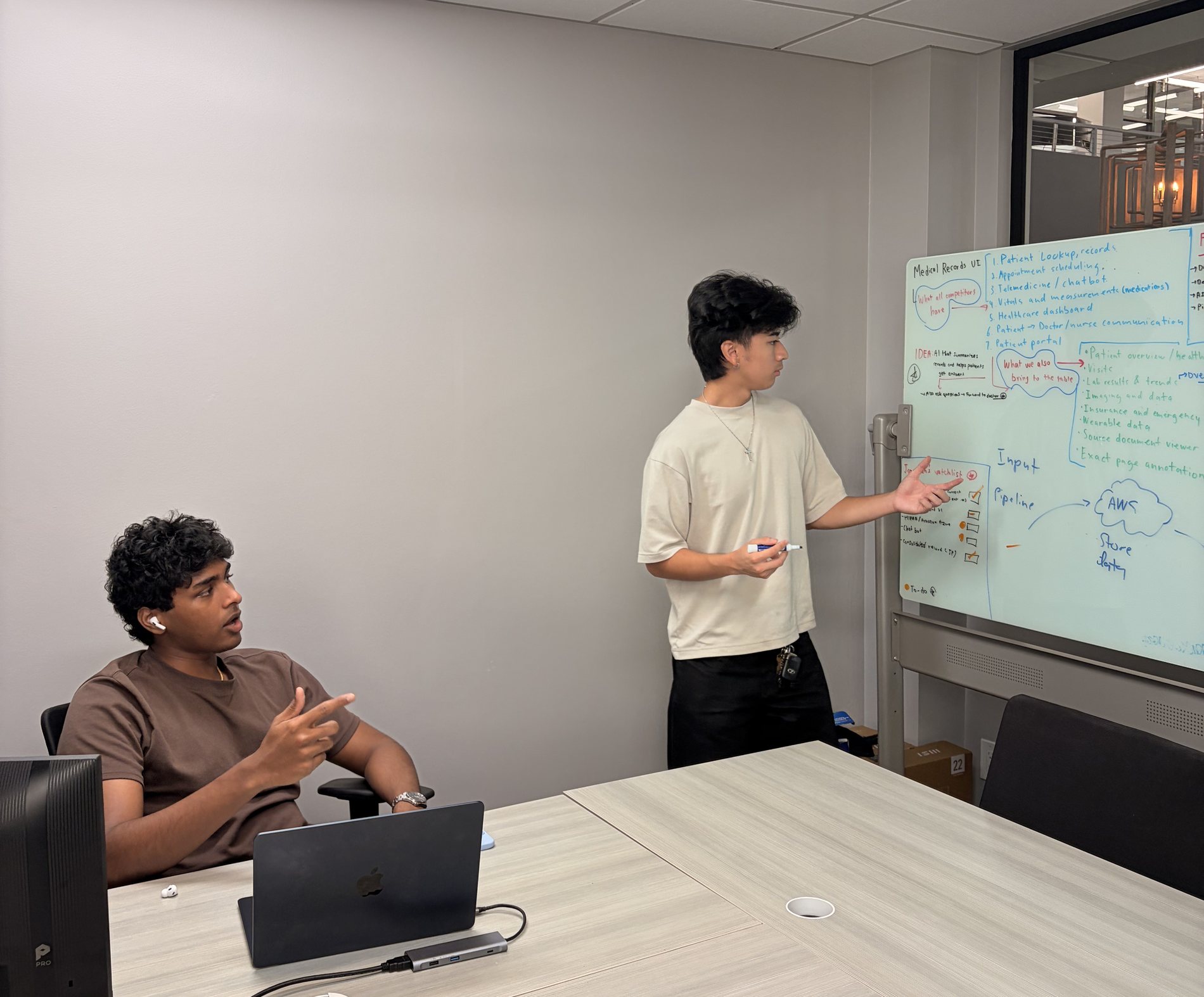 A teammate points to the Medical Records UI plan on the whiteboard while another teammate discusses it from the conference table.